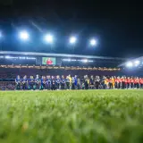 Piłkarze Wisły Kraków i Miedzi Legnica stoją na murawie przed meczem. Stadion wypełniony kibicami, nocne oświetlenie.
