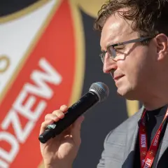 Mężczyzna w okularach z mikrofonem, na tle logo Widzewa. Zapowiada nowe widzew transfery.