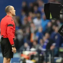 Sędzia w czerwonej koszulce czeka na decyzję VAR. Na boisku trwa mecz, a tłum kibiców obserwuje wydarzenia.