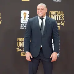 Roberto Carlos żyje i wita fanów na czerwonym dywanie, ubrany w elegancki garnitur. Uśmiecha się szeroko, a w tle widać logo FIFA i napisy związane z Mistrzostwami Świata.