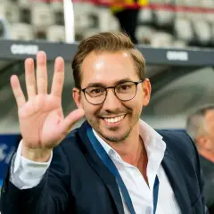 Uśmiechnięty mężczyzna w okularach i garniturze macha ręką na tle trybun stadionu.