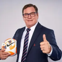Uśmiechnięty trener reprezentacji Polski w garniturze trzyma piłkę i pokazuje kciuk w górę.