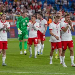 Piłkarze w białych strojach z orzełkiem na piersi, być może przyszli mistrzowie polski w piłce nożnej, stoją na murawie.