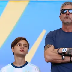 Jorge Messi w okularach przeciwsłonecznych i młody chłopak w koszulce Argentyny na tle niebiesko-żółtego tła.