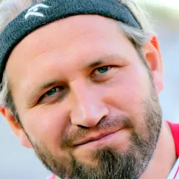 Tomasz Majewski, z opaską na głowie i zarostem, patrzy w obiektyw z lekkim uśmiechem.
