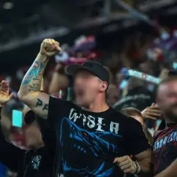 Tatuowany mężczyzna w koszulce z rekinem i napisem "WISŁA" wznosi pięść. W tłumie widać innych kibiców, w tym psycho fans.