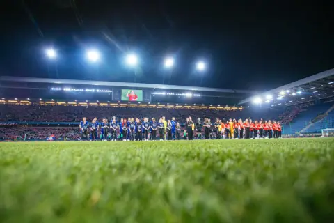 Piłkarze Wisły Kraków i Miedzi Legnica stoją na murawie przed meczem. Stadion wypełniony kibicami, nocne oświetlenie.