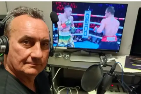 Komentator sportowy w słuchawkach przy mikrofonie, relacjonuje walkę bokserską na ekranie.