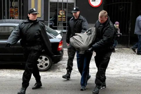Ochroniarze w czarnych kurtkach i czapkach wyprowadzają mężczyznę owiniętego w koc. Może to być interwencja wobec kiboli.