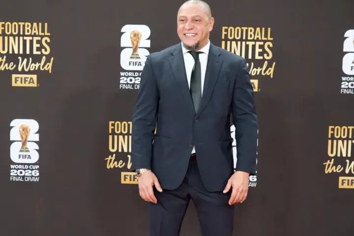 Roberto Carlos żyje i wita fanów na czerwonym dywanie, ubrany w elegancki garnitur. Uśmiecha się szeroko, a w tle widać logo FIFA i napisy związane z Mistrzostwami Świata.