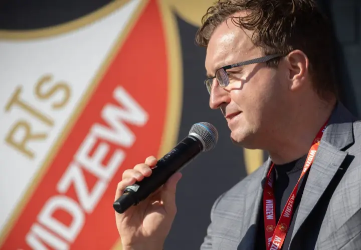 Mężczyzna w okularach z mikrofonem, na tle logo Widzewa. Zapowiada nowe widzew transfery.