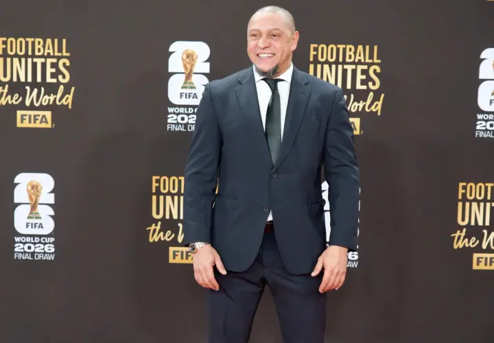 Roberto Carlos żyje i wita fanów na czerwonym dywanie, ubrany w elegancki garnitur. Uśmiecha się szeroko, a w tle widać logo FIFA i napisy związane z Mistrzostwami Świata.
