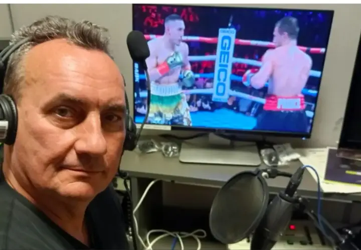 Komentator sportowy w słuchawkach przy mikrofonie, relacjonuje walkę bokserską na ekranie.