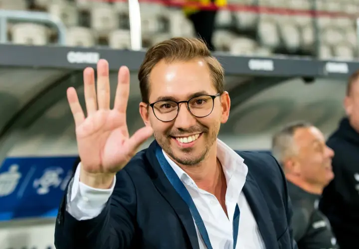Uśmiechnięty mężczyzna w okularach i garniturze macha ręką na tle trybun stadionu.