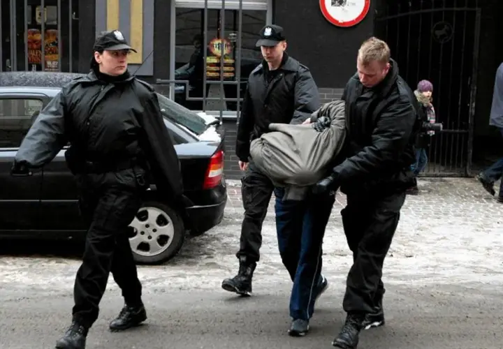 Ochroniarze w czarnych kurtkach i czapkach wyprowadzają mężczyznę owiniętego w koc. Może to być interwencja wobec kiboli.