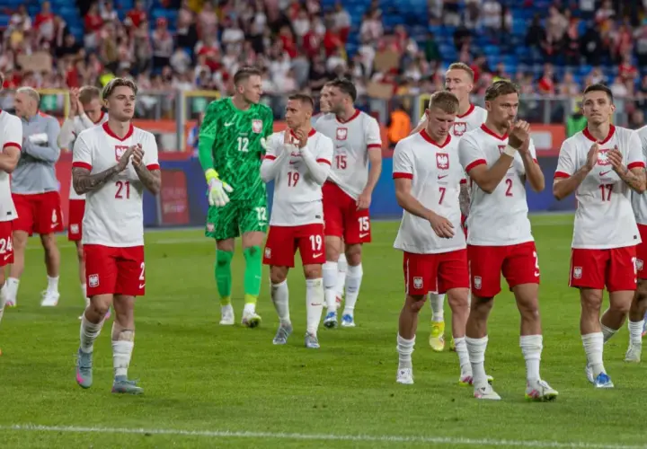 Piłkarze w białych strojach z orzełkiem na piersi, być może przyszli mistrzowie polski w piłce nożnej, stoją na murawie.