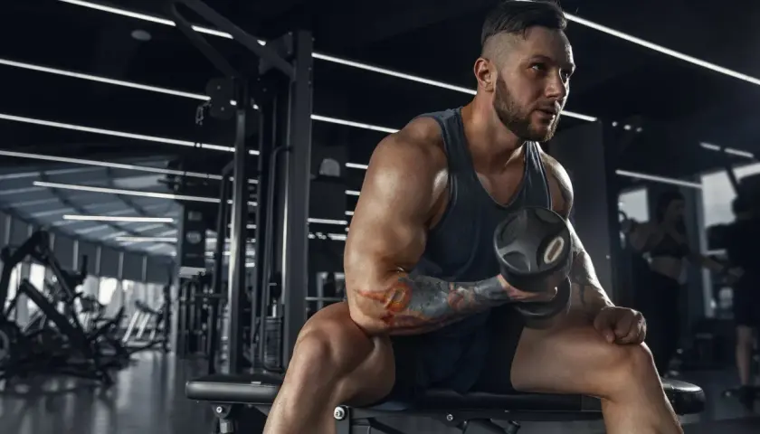 Muskularny mężczyzna z tatuażami ćwiczy biceps z hantlem w siłowni. Jego determinacja jest widoczna, jakby każdy ruch przybliżał go do celu, być może wspomaganego przez clenbuterol.