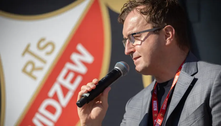 Mężczyzna w okularach z mikrofonem, na tle logo Widzewa. Zapowiada nowe widzew transfery.
