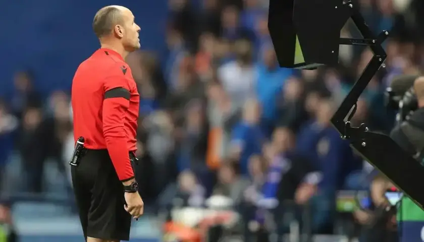 Sędzia w czerwonej koszulce czeka na decyzję VAR. Na boisku trwa mecz, a tłum kibiców obserwuje wydarzenia.