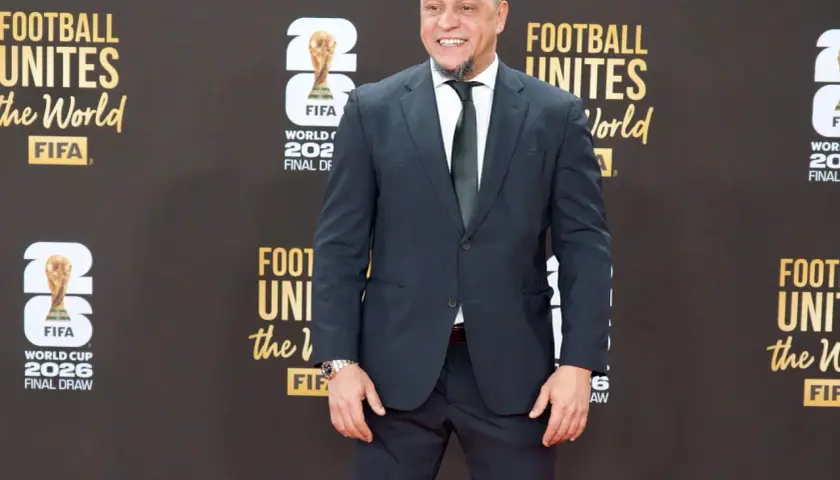 Roberto Carlos żyje i wita fanów na czerwonym dywanie, ubrany w elegancki garnitur. Uśmiecha się szeroko, a w tle widać logo FIFA i napisy związane z Mistrzostwami Świata.