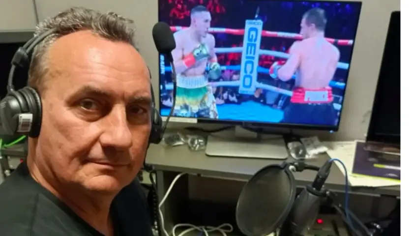 Komentator sportowy w słuchawkach przy mikrofonie, relacjonuje walkę bokserską na ekranie.