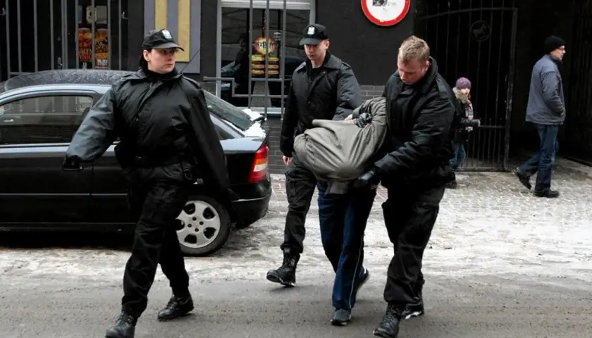 Ochroniarze w czarnych kurtkach i czapkach wyprowadzają mężczyznę owiniętego w koc. Może to być interwencja wobec kiboli.