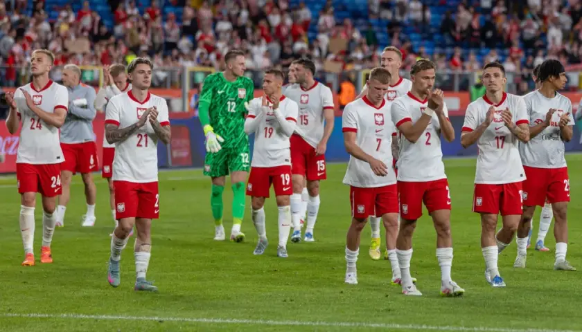 Piłkarze w białych strojach z orzełkiem na piersi, być może przyszli mistrzowie polski w piłce nożnej, stoją na murawie.