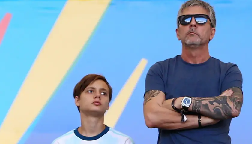 Jorge Messi w okularach przeciwsłonecznych i młody chłopak w koszulce Argentyny na tle niebiesko-żółtego tła.