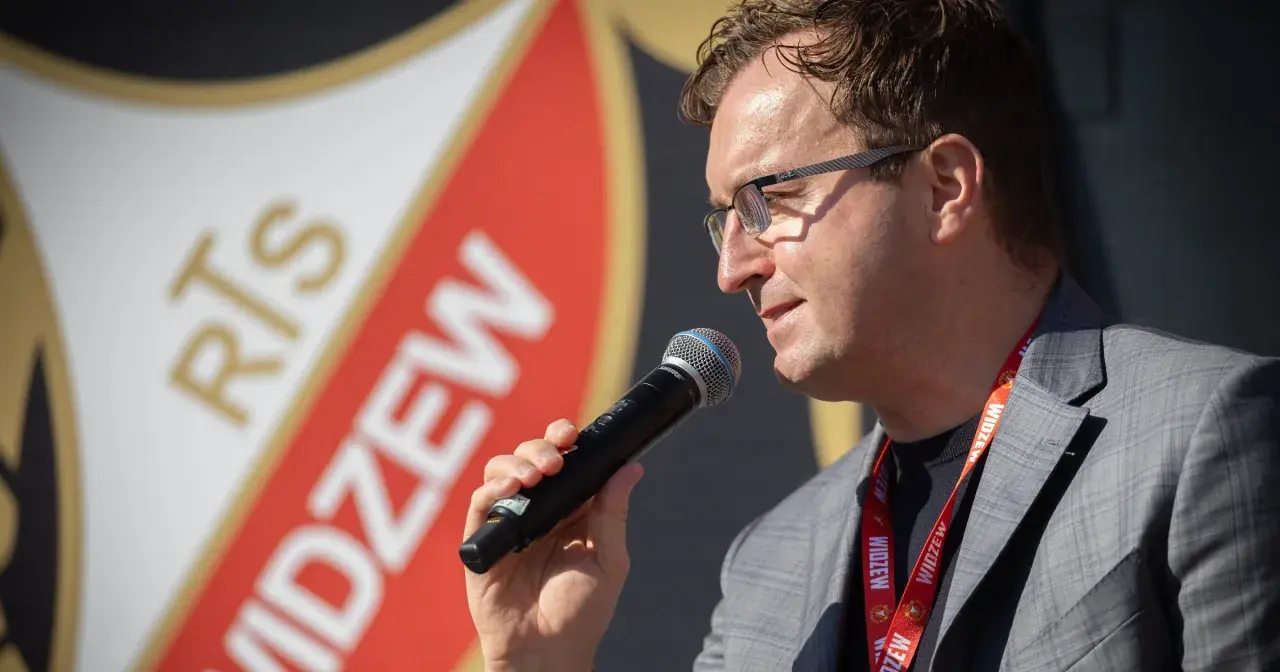 Mężczyzna w okularach z mikrofonem, na tle logo Widzewa. Zapowiada nowe widzew transfery.
