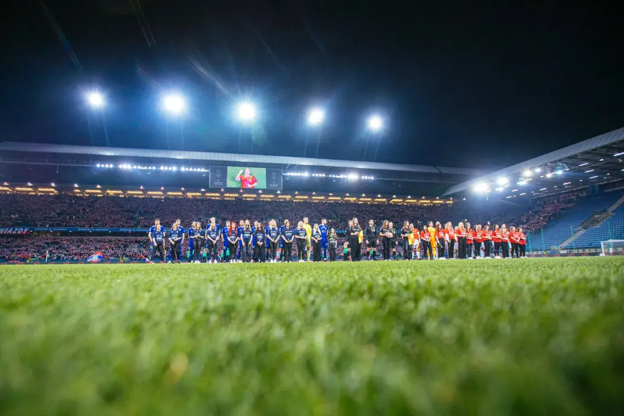 Piłkarze Wisły Kraków i Miedzi Legnica stoją na murawie przed meczem. Stadion wypełniony kibicami, nocne oświetlenie.