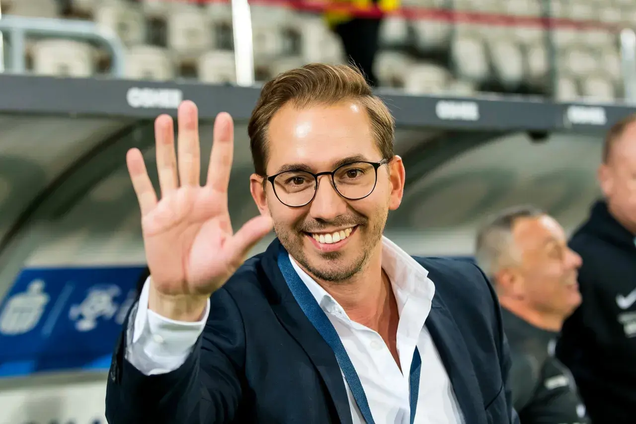 Uśmiechnięty mężczyzna w okularach i garniturze macha ręką na tle trybun stadionu.