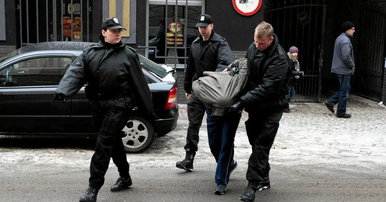 Ochroniarze w czarnych kurtkach i czapkach wyprowadzają mężczyznę owiniętego w koc. Może to być interwencja wobec kiboli.