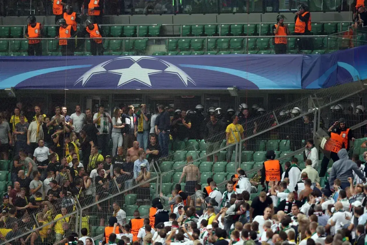 Tłum kibiców na stadionie, wśród nich widać ochronę i policję. Niektórzy kibice wyglądają na zdenerwowanych, co może sugerować napiętą atmosferę.