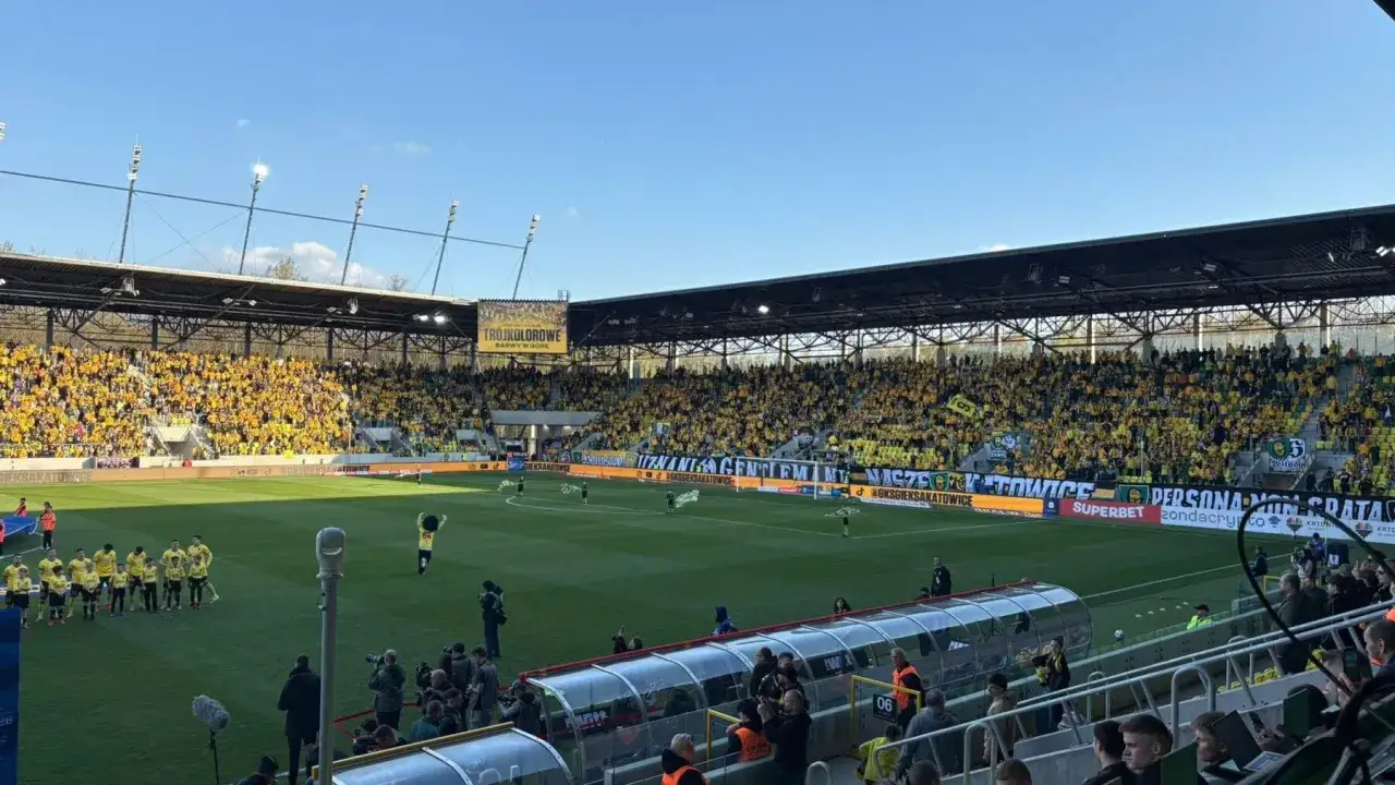 Stadion wypełniony kibicami GKS Katowice, którzy dopingują swoją drużynę. W tle transparenty z nazwami klubu, a na boisku zawodnicy.