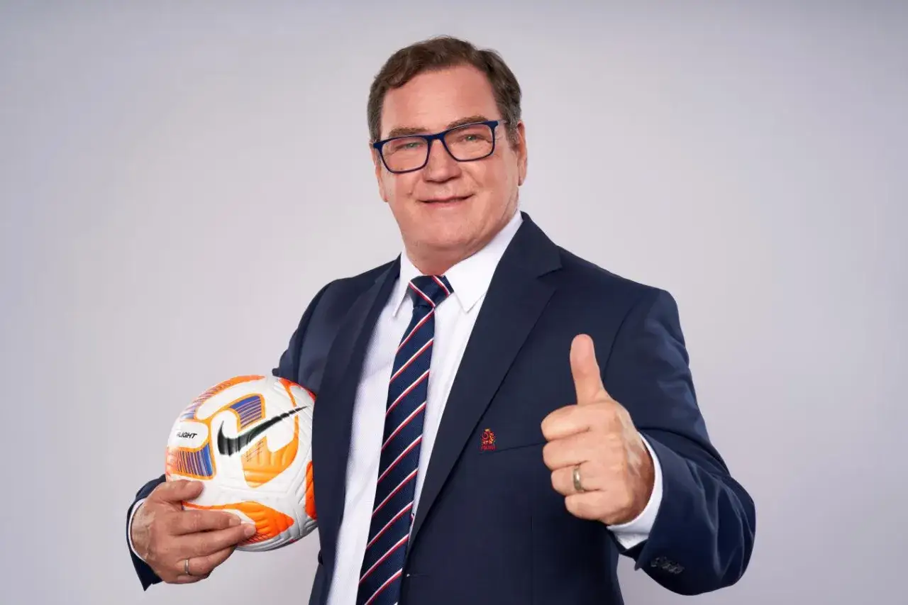 Uśmiechnięty trener reprezentacji Polski w garniturze, z piłką w ręku i kciukiem w górze.