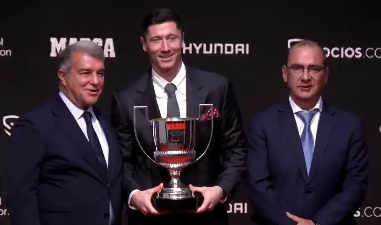 Robert Lewandowski z pucharem Pichichi, uśmiechnięty, w otoczeniu dwóch mężczyzn w garniturach.