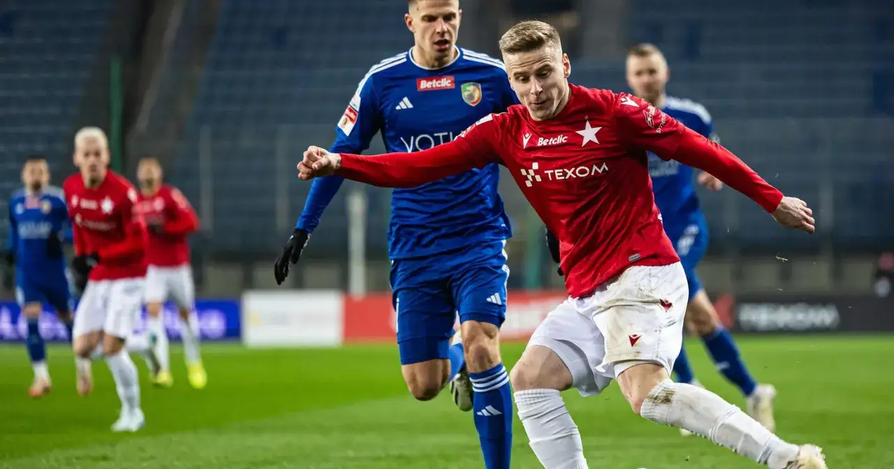 Piłkarz Wisły Kraków w akcji podczas meczu z Miedzią Legnica. Zawodnik w czerwonej koszulce biegnie z piłką, ścigany przez rywala w niebieskim stroju.