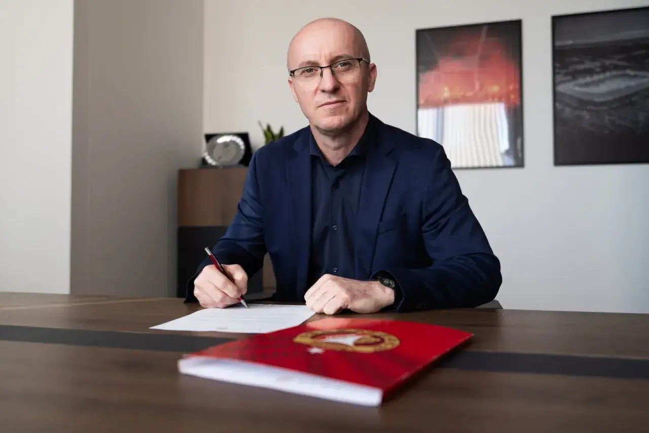 Nowy trener Widzewa podpisuje kontrakt. Mężczyzna w okularach siedzi przy biurku, pisząc piórem.