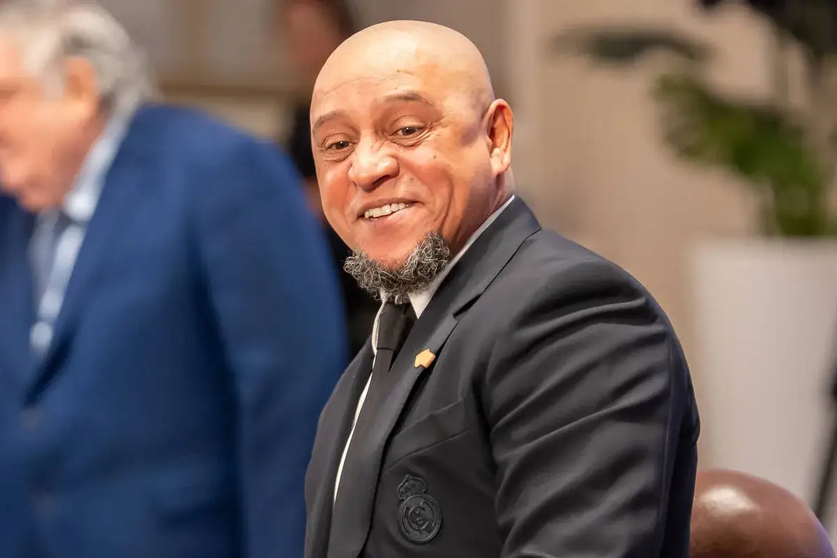 Uśmiechnięty Roberto Carlos w garniturze, z charakterystyczną brodą. Widać herb Realu Madryt. Roberto Carlos żyje i ma się dobrze!