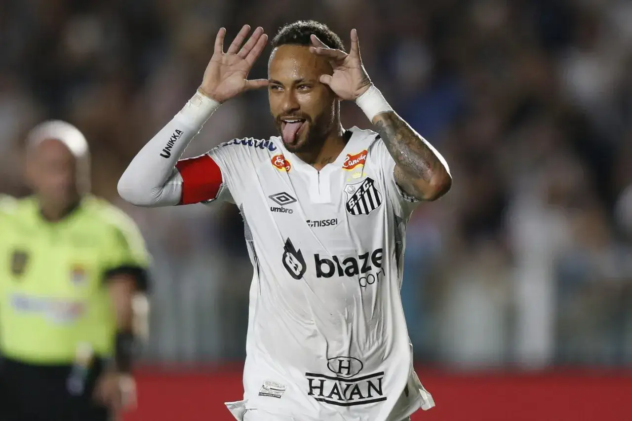 Neymar w białej koszulce Santos, z językiem na wierzchu, pokazuje radość z gry.